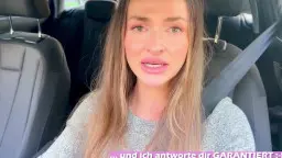 Brünette in grauem Kleid sitzt angeschnallt am Steuer und blickt den Betrachter direkt an.