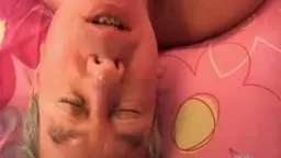 Nahaufnahme eines älteren Mannes, der mit geschlossenen Augen und offenem Mund lustvoll in eine pinke Bettwäsche gedrückt wird.