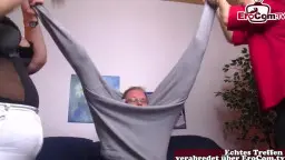 Älterer Mann liegt mit dem Kopf zwischen den Beinen der beiden Blondinen auf dem Sofa, während sie ihm ihre nackten Körper präsentieren