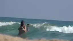 Nackte Brünette mit Zöpfen steht mit dem Rücken zur Kamera in der Brandung und filmt sich mit dem Handy beim Baden im Meer.
