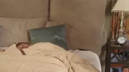 Meli steht verführerisch in weißem Top und Short neben dem Bett, während ein Mann darunter schläft.