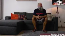 Mollige blonde Oma im roten Kleid sitzt auf grauem Sofa neben älterem Mann mit weißem Bart