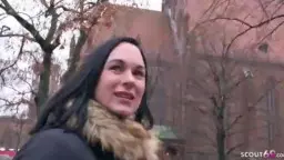 Brünette Milf mit Pelzkragenjacke lächelt frech vor alter Backsteinkirche